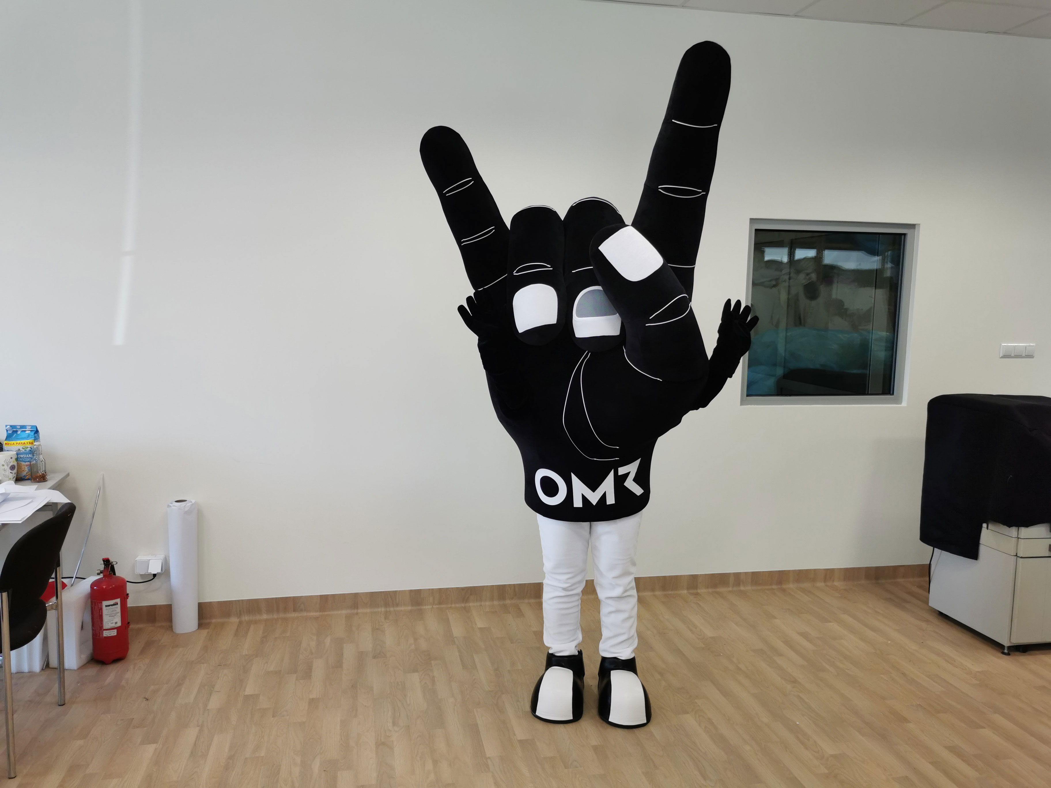 Na fotografii widnieje maskotka w postaci stroju reklamowego stylizowanego na dłoń o anatomicznym wyglądzie. Ręka w kolorze czarnym pozuje do zdjęcia w siedzibie firmy Elabika